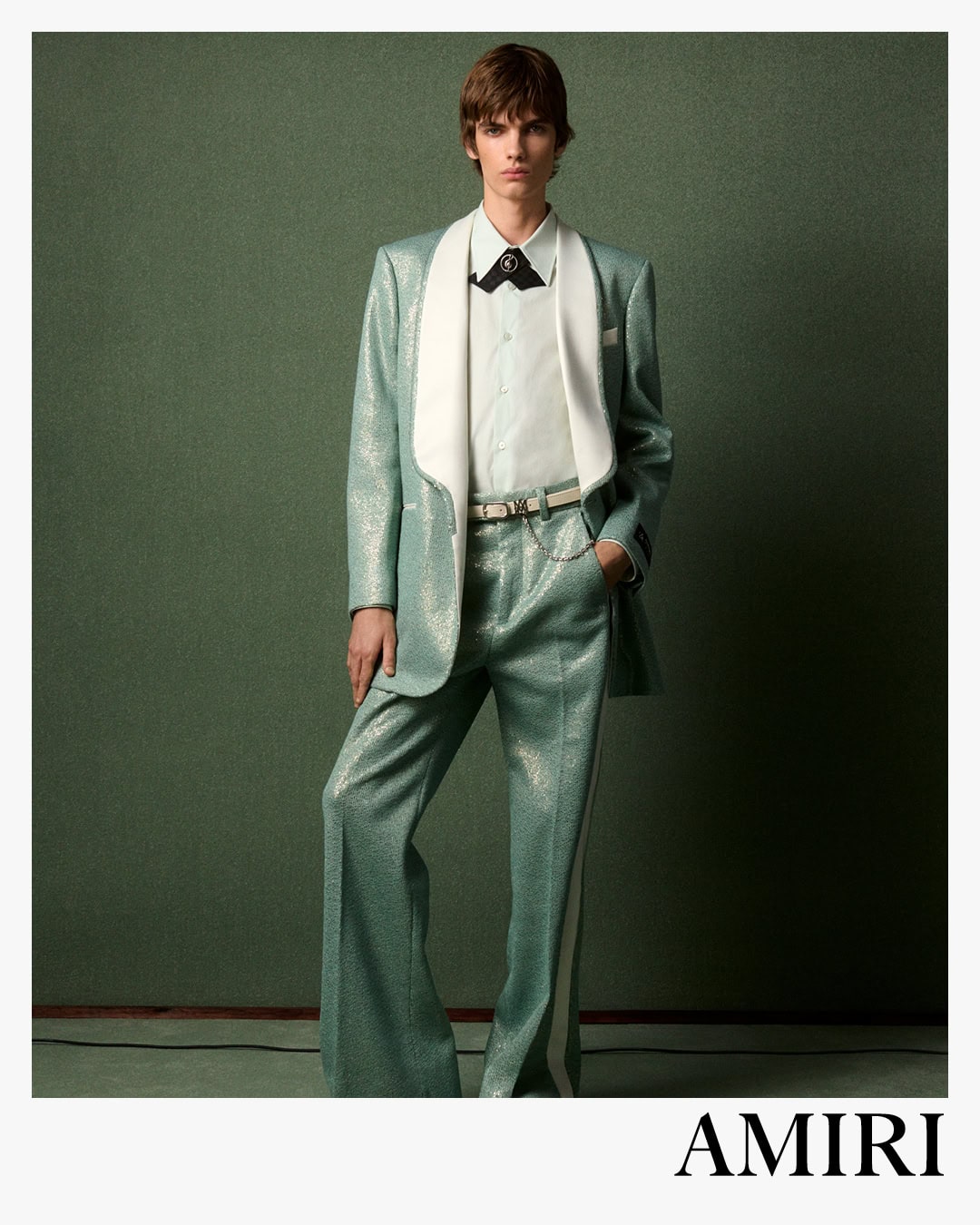 Model wearing a metallic sage green suit with white lapels and a white shirt against a textured green background