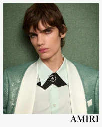 Portrait of a male model wearing a mint green jacket, white collared shirt, and black necktie with silver accessory against textured green background