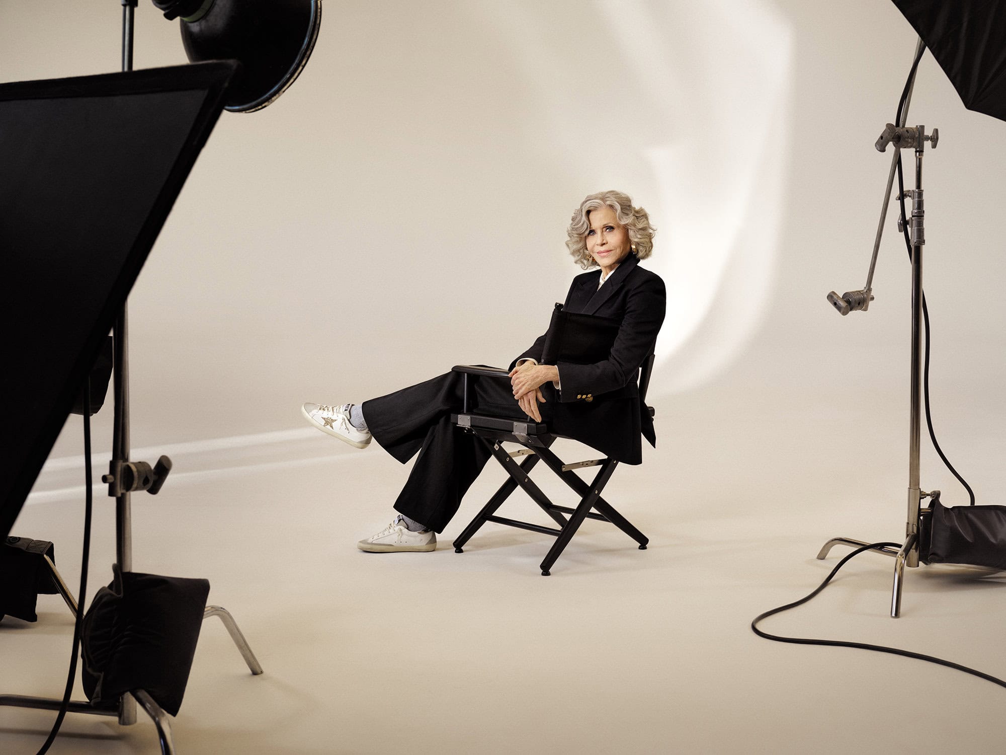 Jane Fonda seated in a director chair wearing a black suit and sneakers in a professional photo studio setting