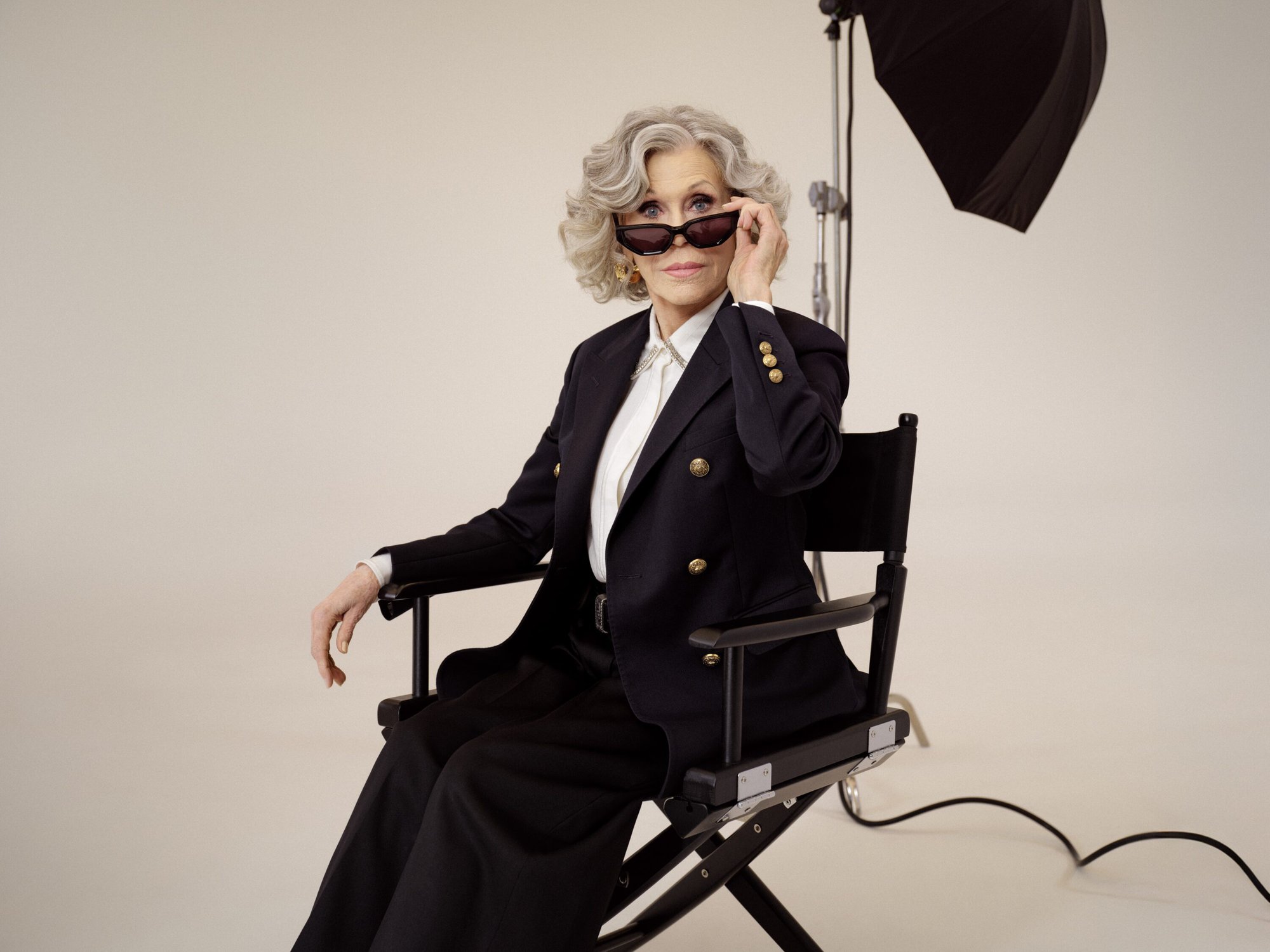 Jane Fonda seated in a director chair wearing dark sunglasses and a black blazer against a white seamless background.