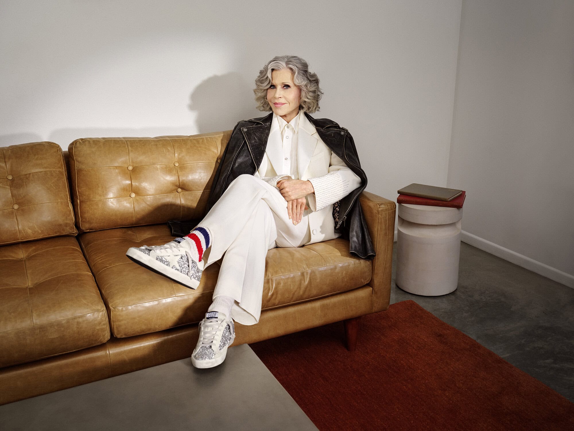 Jane Fonda seated on a tan leather couch wearing a white suit, black leather jacket, and patterned sneakers