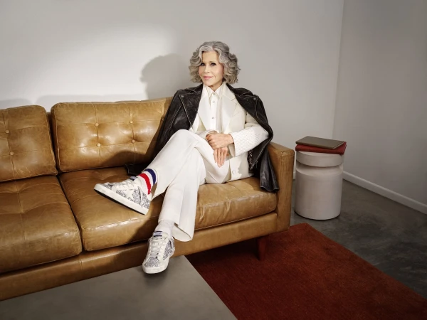 Jane Fonda seated on a tan leather couch wearing a white suit, black leather jacket, and patterned sneakers
