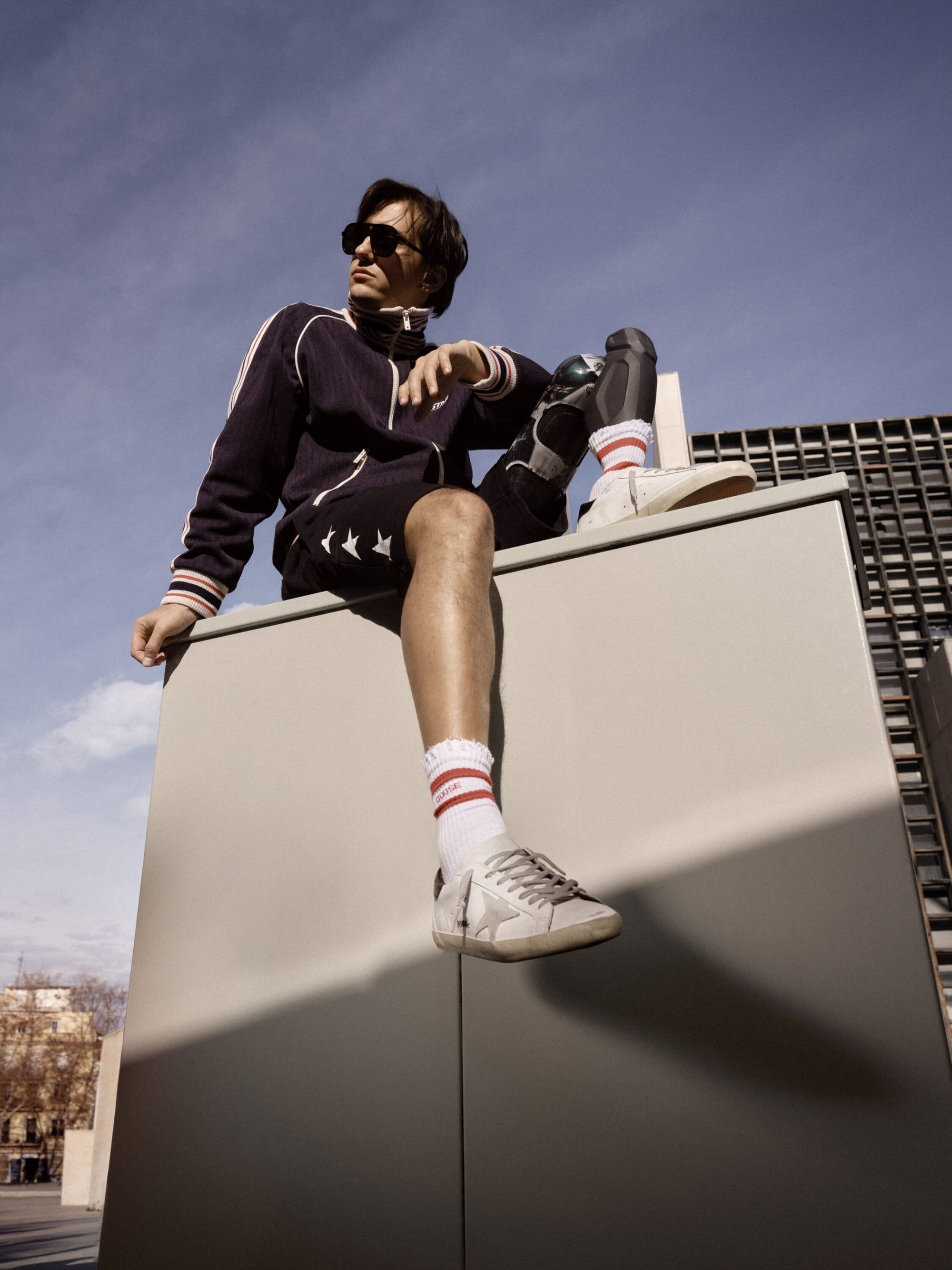 Man wearing sunglasses and athletic wear sitting atop a concrete structure against a blue sky