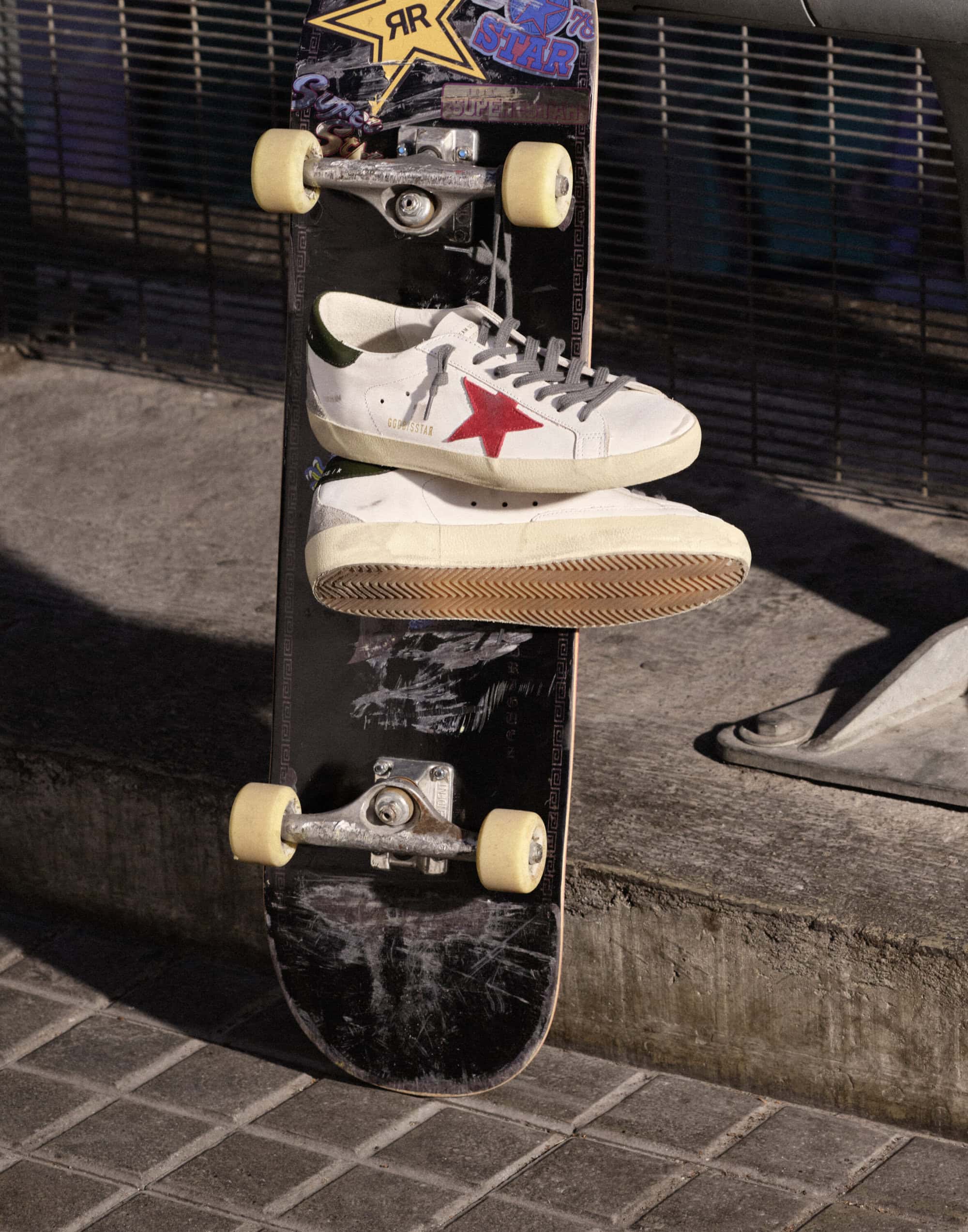 White leather sneakers with red star resting on a black skateboard propped against an urban metal fence.