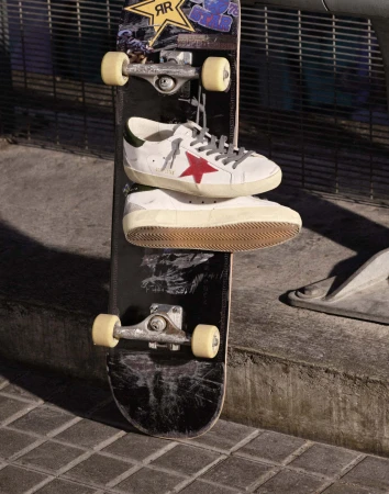 White leather sneakers with red star resting on a black skateboard propped against an urban metal fence.