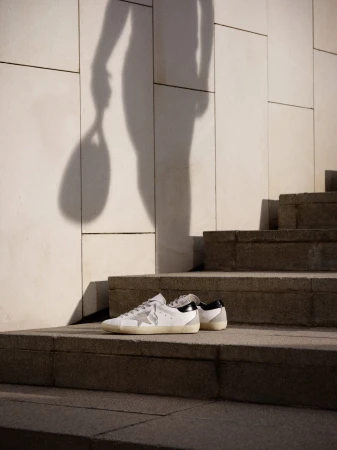 Golden Goose sneakers rest on stone stairs as a person's shadow holding a racket is cast on a tiled wall behind them.