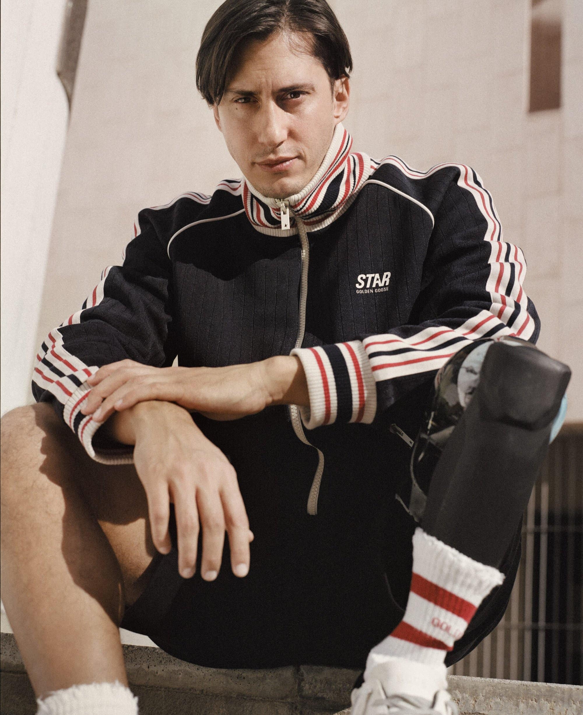 Man wearing a black track jacket with red and white stripes and a prosthetic leg posing against an architectural building exterior.
