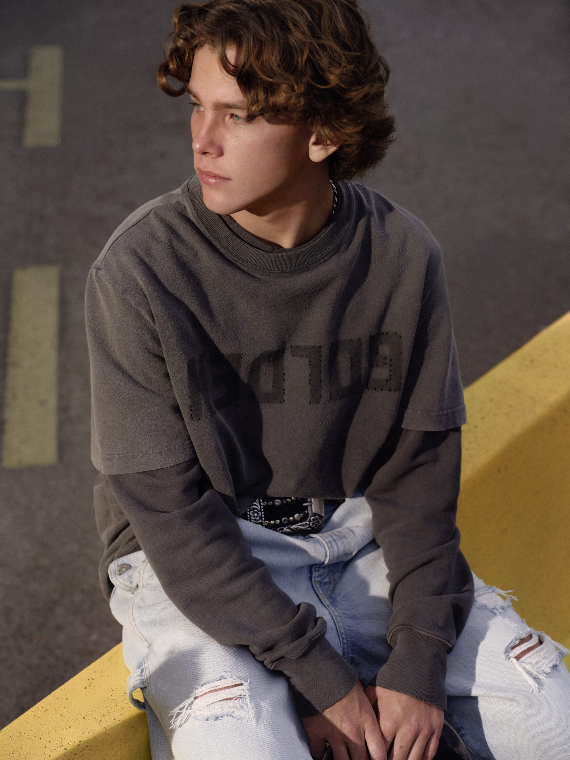 Young man with curly brown hair wearing a dark grey sweatshirt and light wash distressed denim jeans outdoors