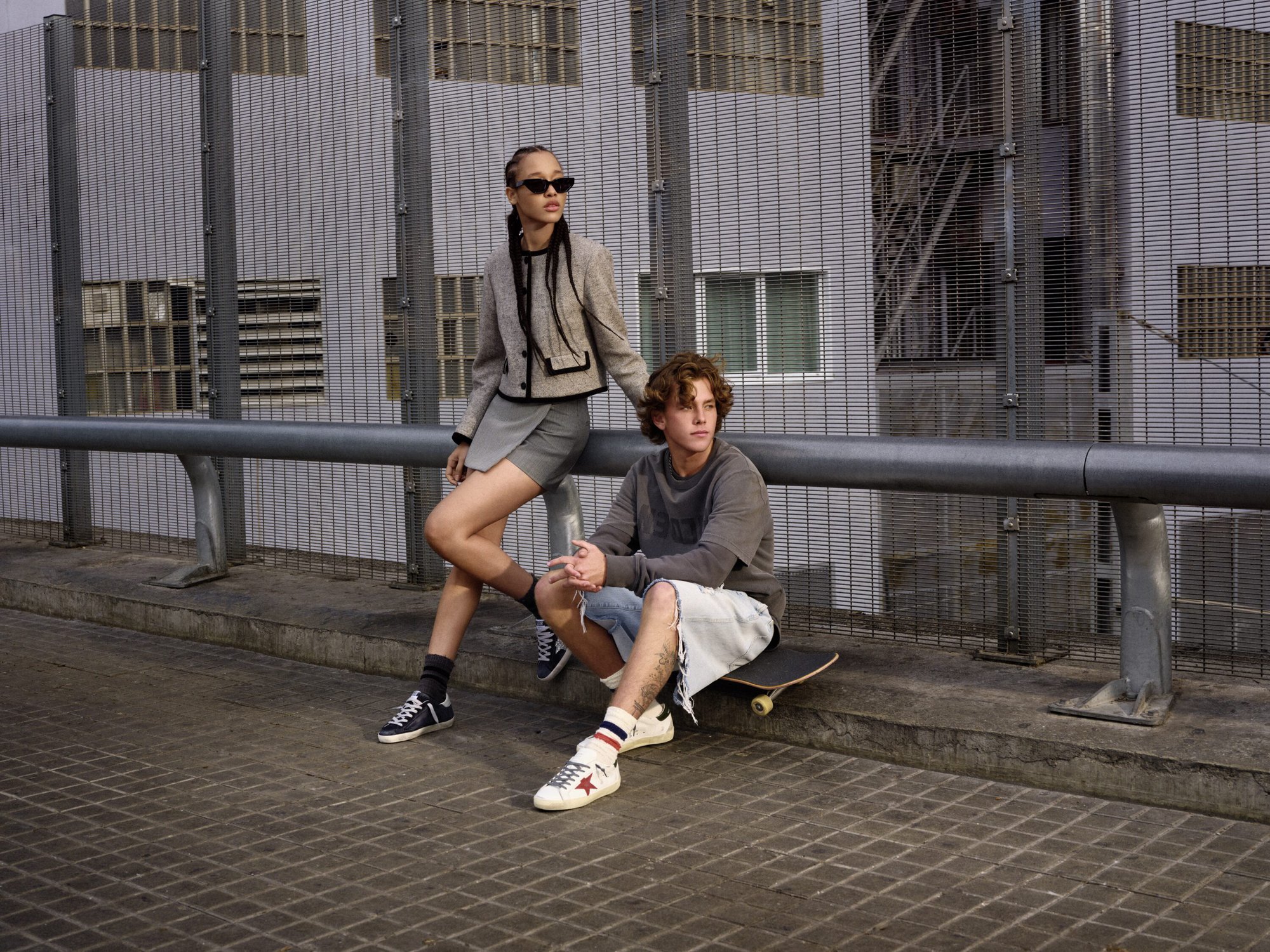 Two models posing outdoors against a metal security fence and concrete structure for Golden Goose campaign