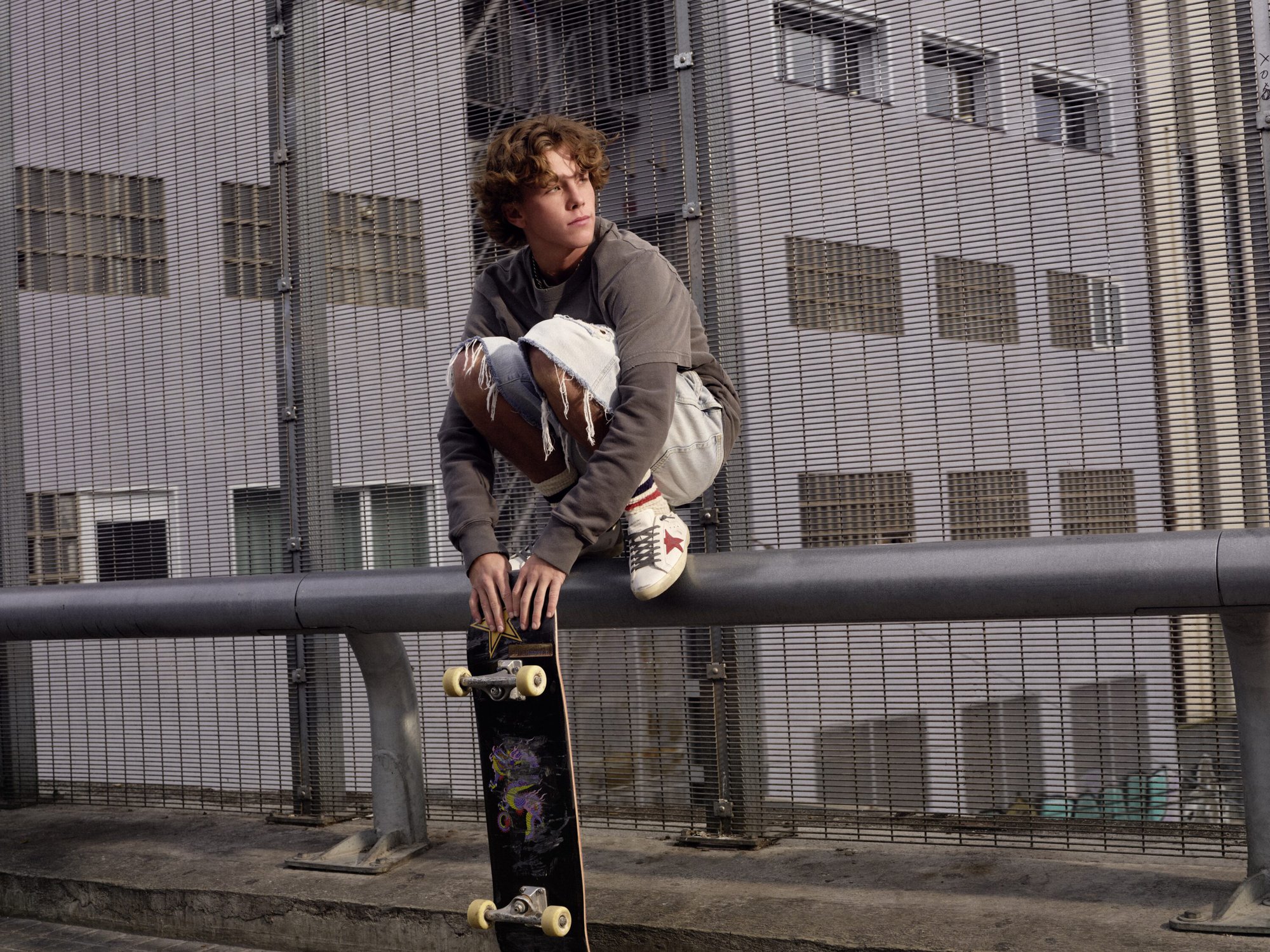 Young man crouching on a metal railing outdoors with a skateboard, wearing casual streetwear against a building facade.