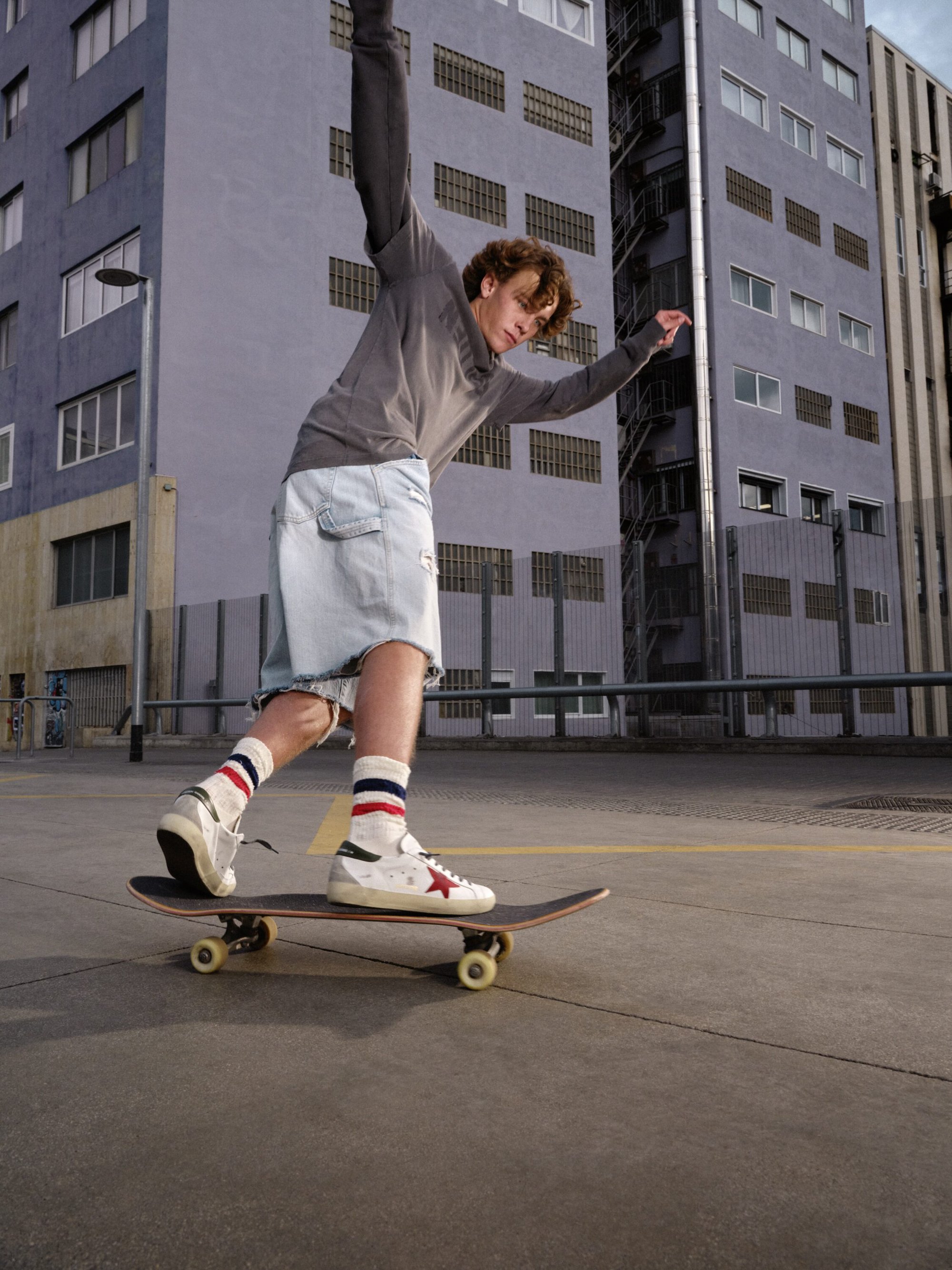 Young man skateboarding outdoors on a paved surface wearing loose shorts and sneakers, with tall grey buildings in the background.