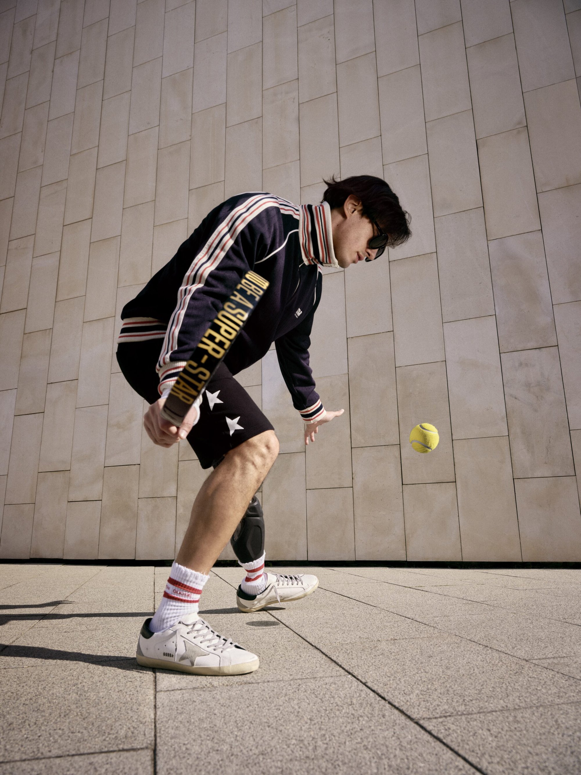 Man wearing dark athletic track jacket and shorts bouncing tennis ball on paddle against stone wall background