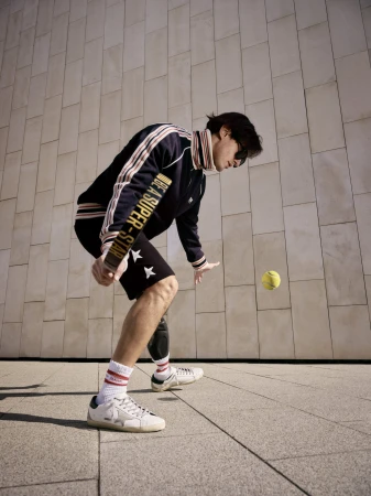 Man wearing dark athletic track jacket and shorts bouncing tennis ball on paddle against stone wall background