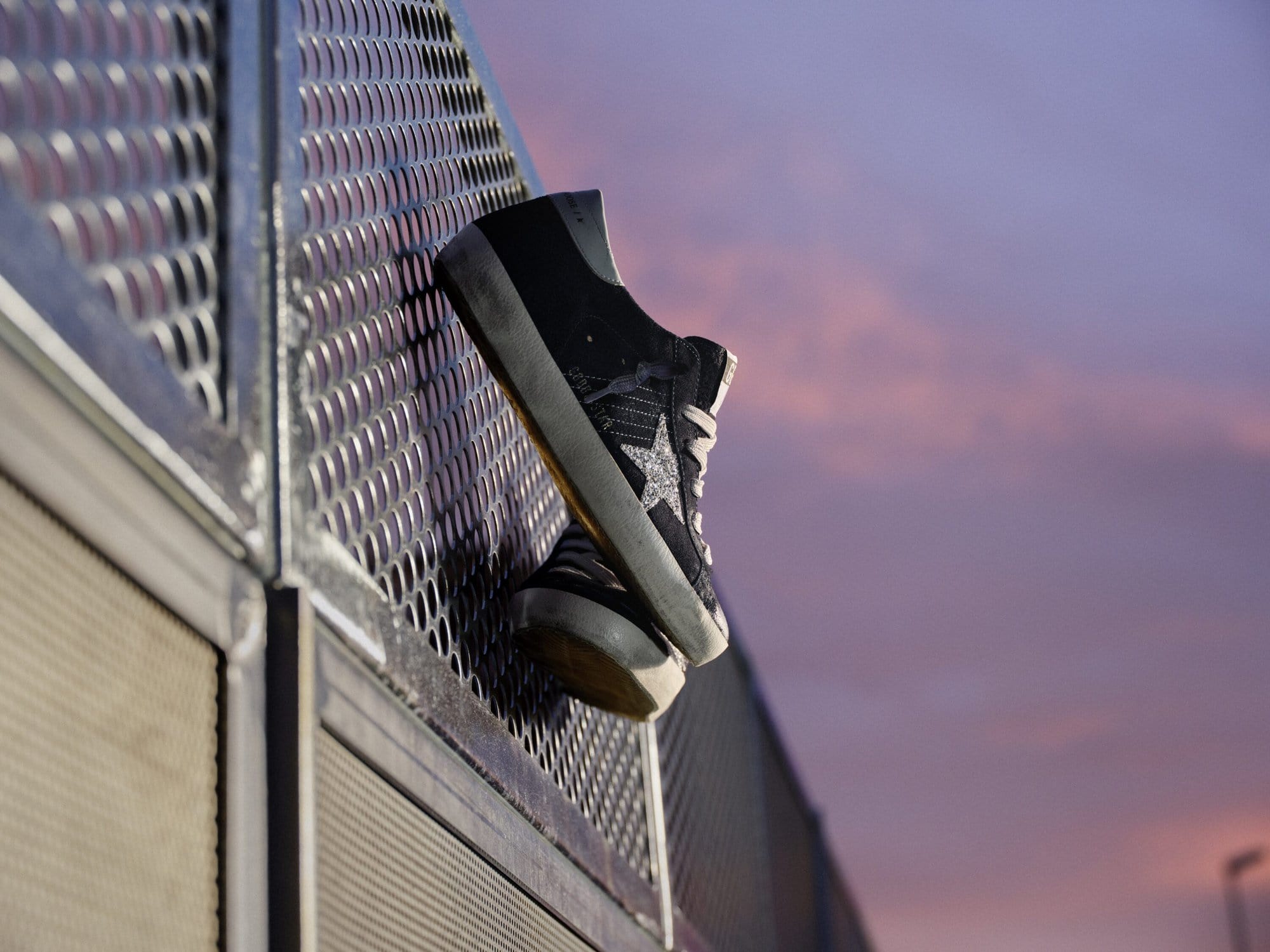 Black and glitter sneaker placed against a perforated metal barrier at dusk