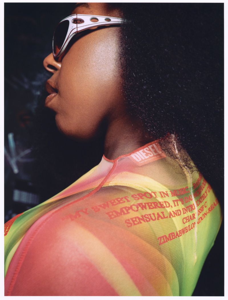 Woman with dark hair, wearing stylish sunglasses and a colorful transparent top with text. Profile portrait against a dark background.