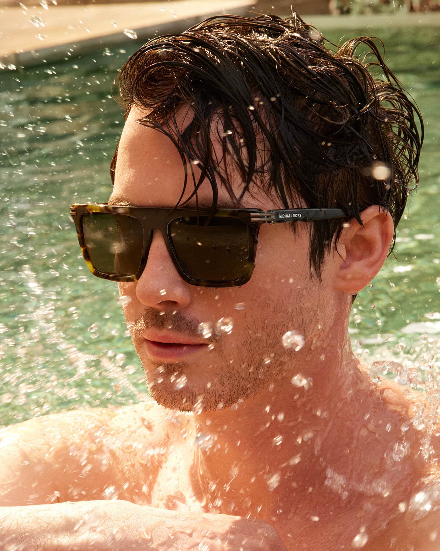 Logan Lerman wearing tortoiseshell sunglasses in an outdoor pool with water splashing around him