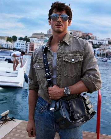 Logan Lerman in aviator sunglasses wearing an olive jacket and blue jeans by a marina in Ibiza for Michael Kors Spring 2025 ad campaign.