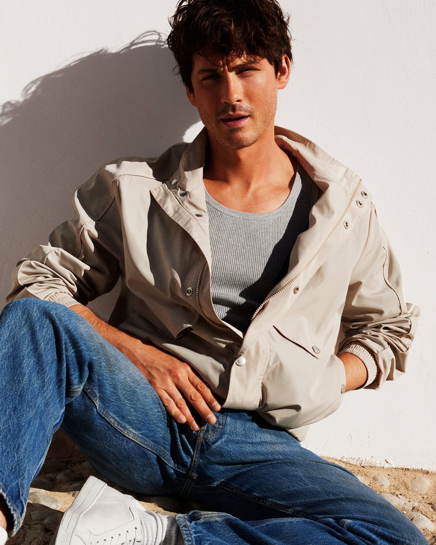 Man wearing a beige jacket and blue jeans seated against a bright white wall for Michael Kors spring campaign