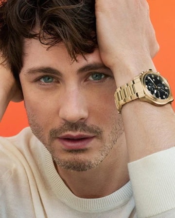 Close-up portrait of Logan Lerman wearing a gold watch and white sweater against an orange background