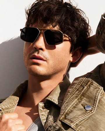 Close-up portrait of a man wearing dark aviator sunglasses and an olive green jacket against a bright white wall.