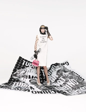Model in white dress standing on printed paper sheets holding a pink handbag for Moschino Spring 2025 ad campaign.