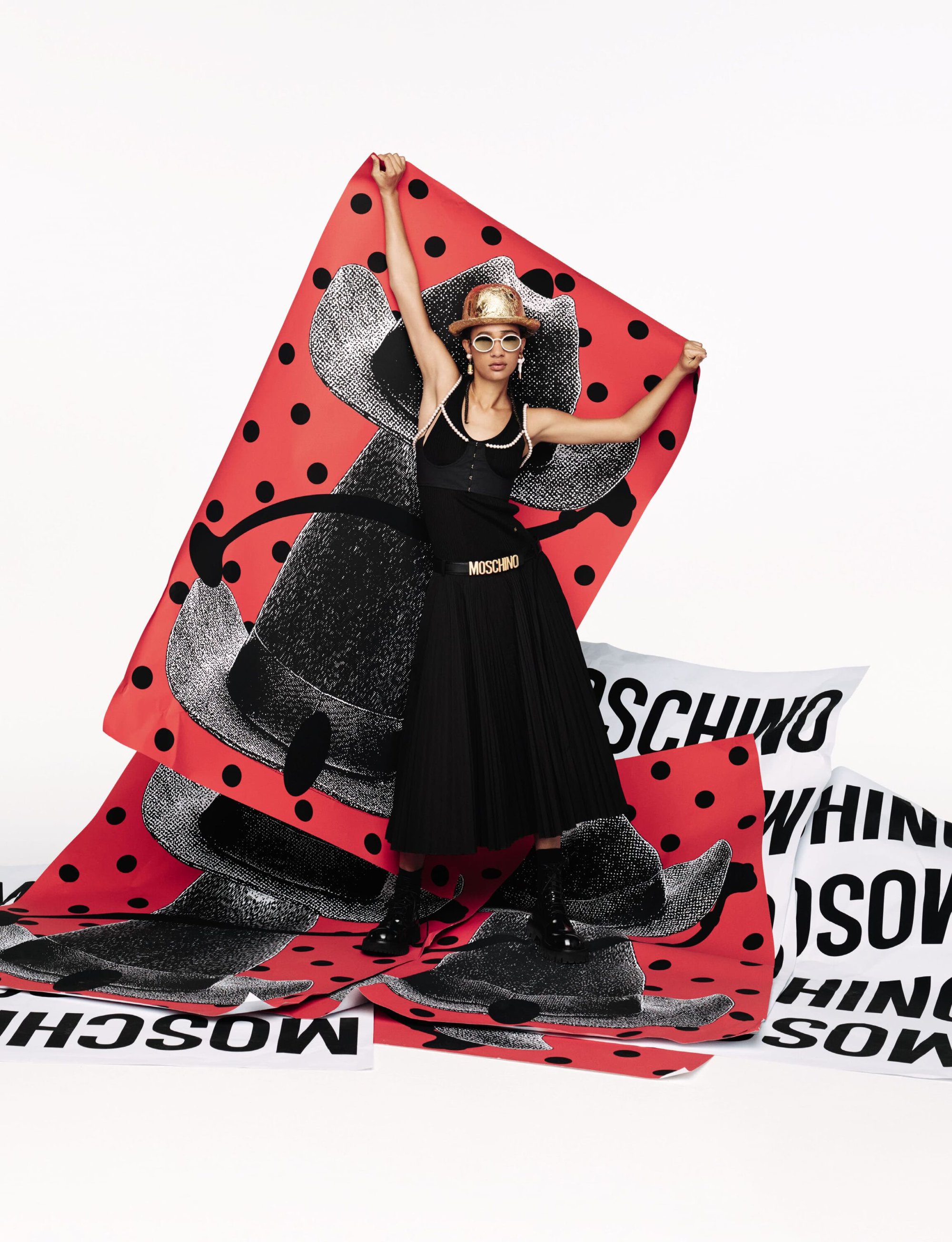 Model in black dress holding a large red patterned backdrop featuring a top hat graphic for Moschino Spring 2025 campaign.