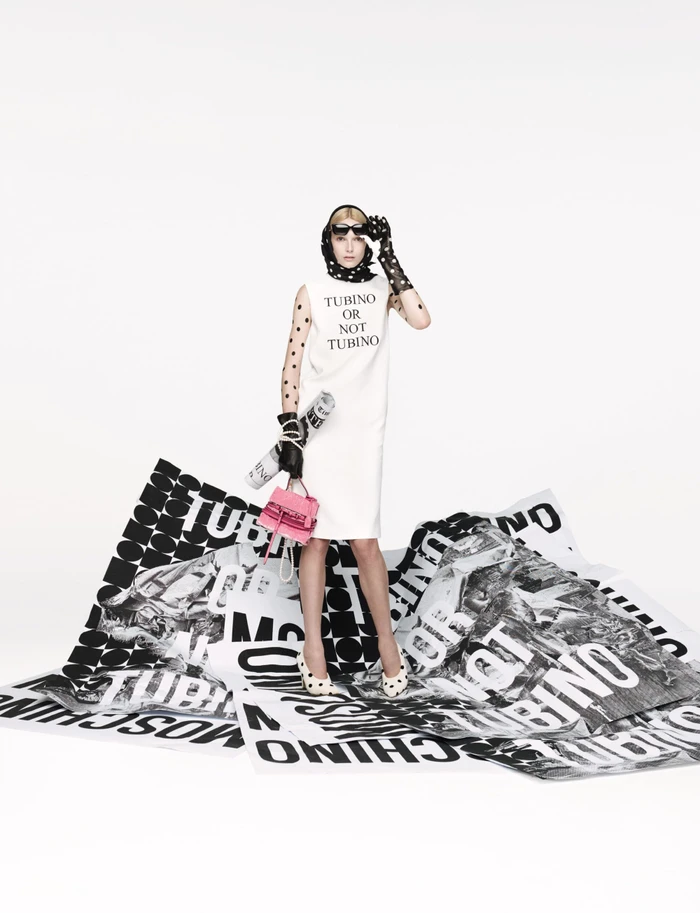 Model in white dress standing on printed paper sheets holding a pink handbag for Moschino Spring 2025 ad campaign.