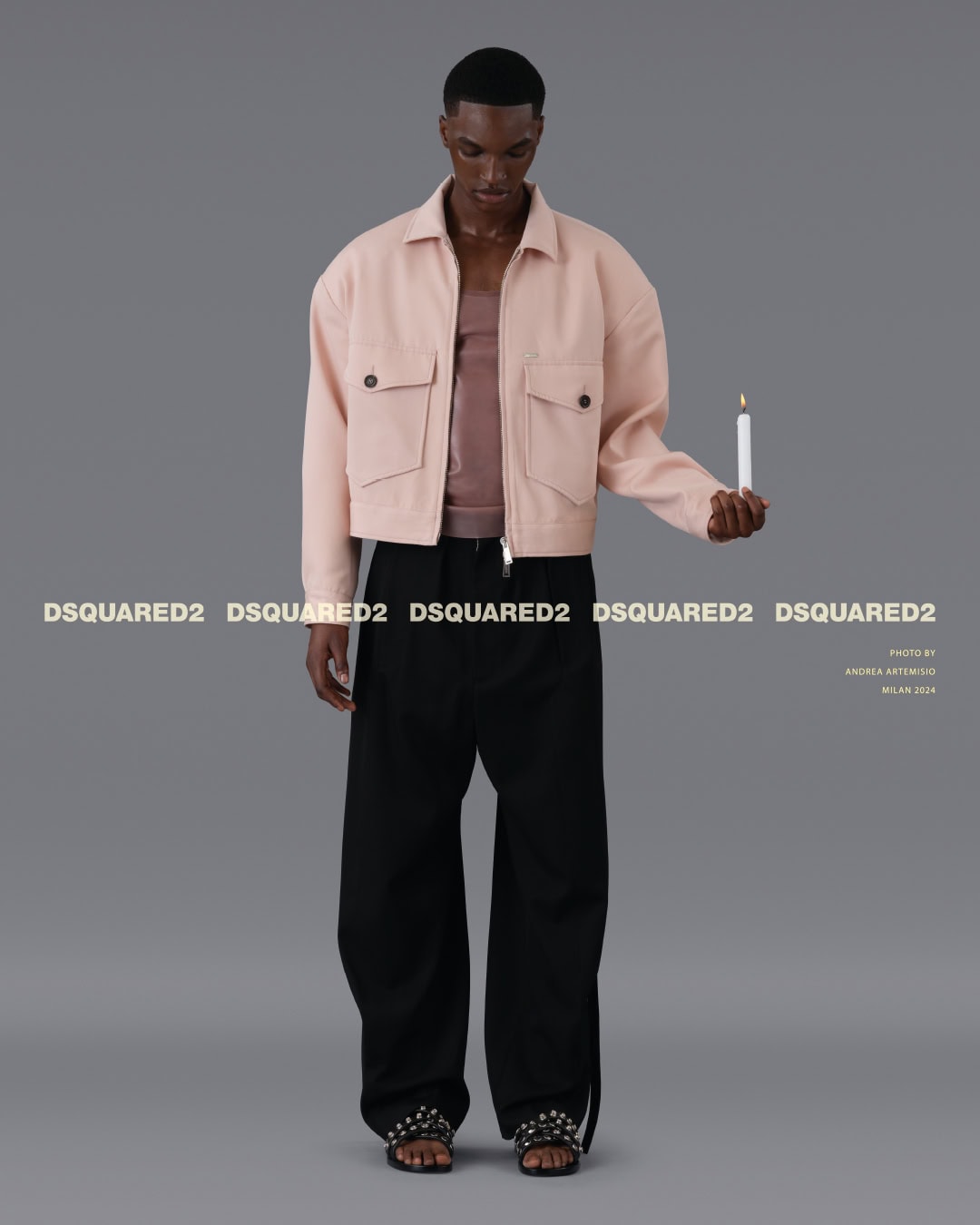 Man wearing a light pink jacket and black trousers holding a lit candle against a grey studio backdrop