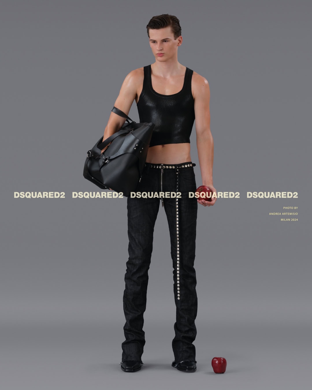 A young male model in a black tank top and dark jeans holding a leather duffel bag against a grey seamless studio background.