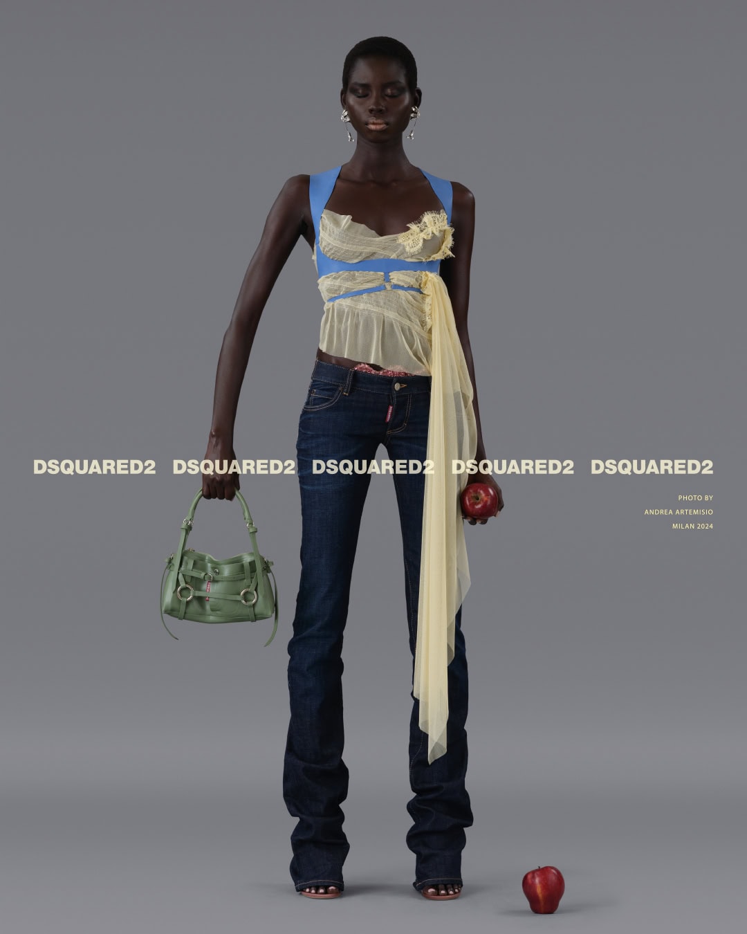 Black model wearing a layered sheer top and dark denim jeans against a plain grey backdrop holding a green handbag