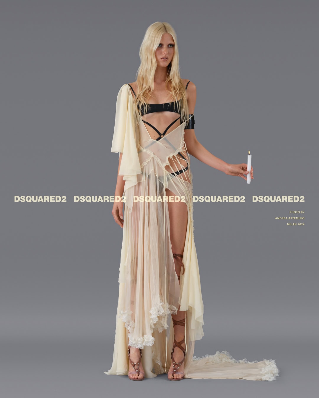 Model with long blonde hair wearing a sheer layered cream dress holding a lit candle against a grey studio backdrop