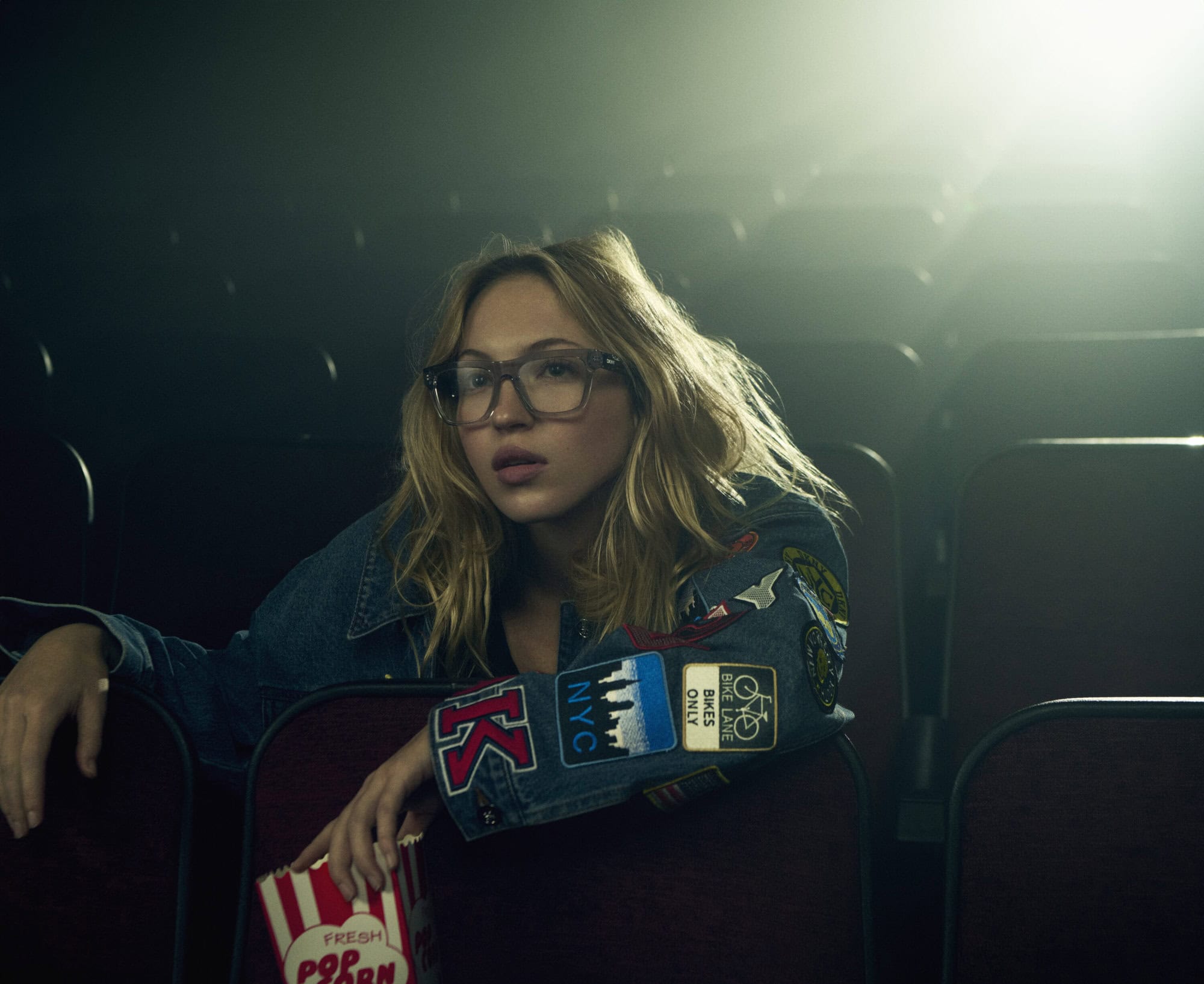 Model Lila Moss seated in a dark cinema holding a popcorn carton wearing patches on a denim jacket for DKNY Spring 2025 Ad Campaign.