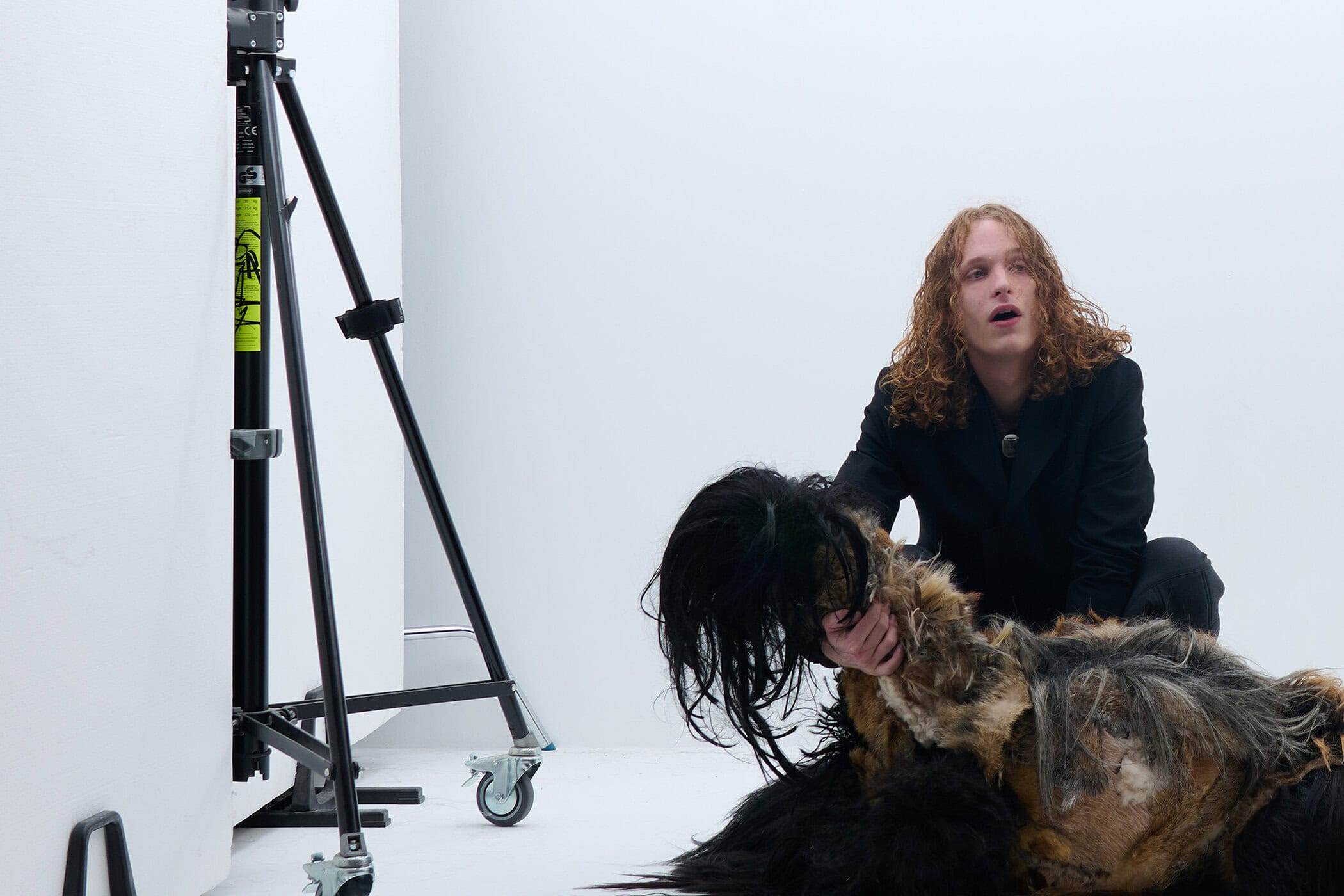 A man with long curly red hair in a black blazer crouches next to a shaggy dark prop in a white studio.