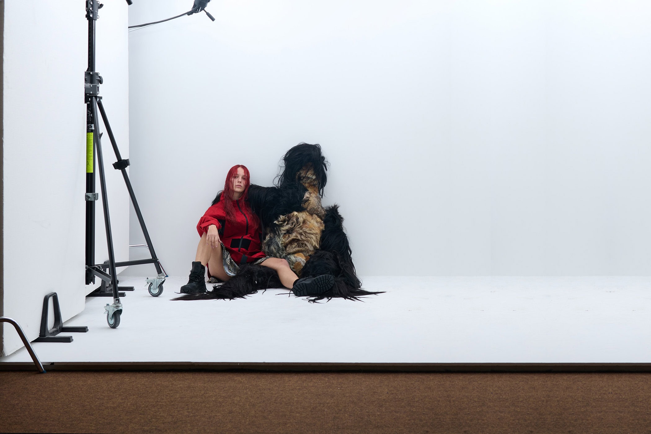 Model with red hair wearing a red jacket and boots seated next to a shaggy black fur sculpture in a white studio