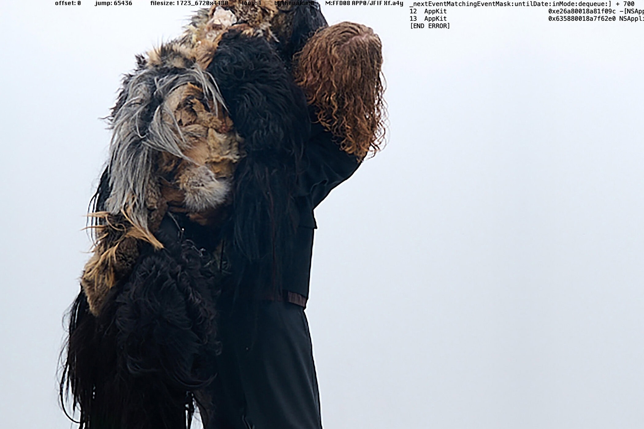 Person with long curly hair wearing dark clothing carrying a bulky textured garment against an overcast sky