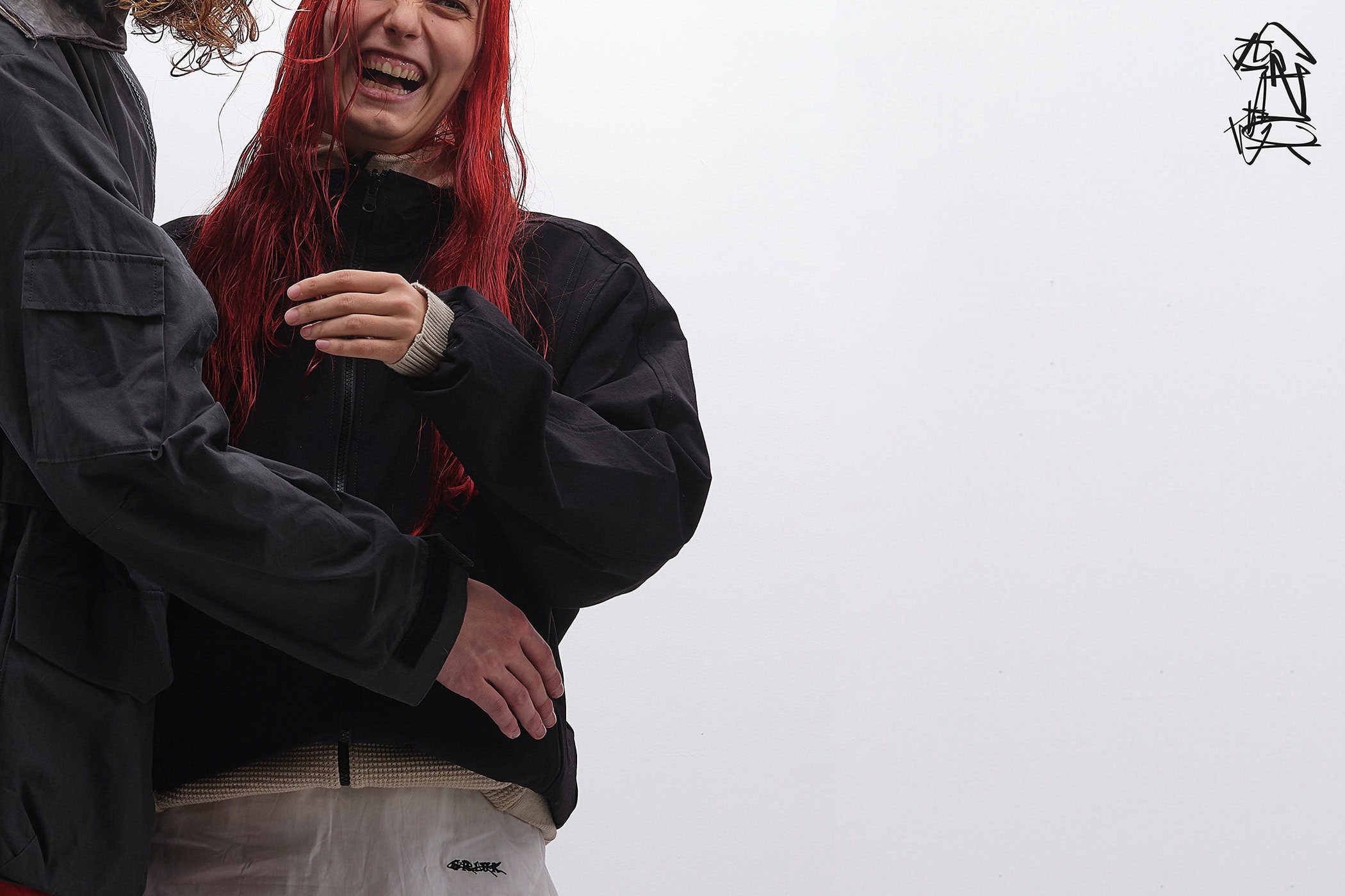 Close-up of a smiling woman with red hair wearing a black jacket and white pants standing beside another person against a plain white background.