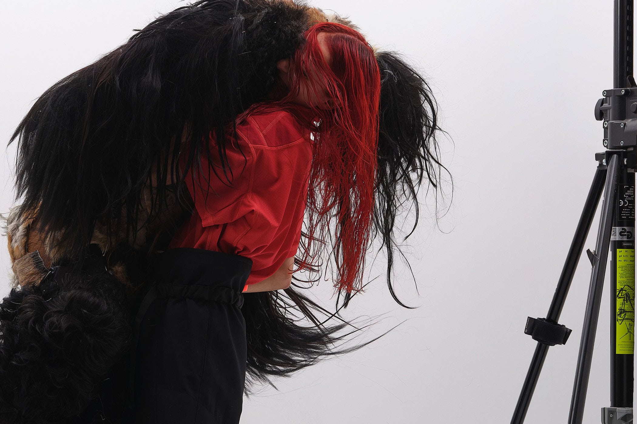 Person with long red hair wearing a red top and black trousers embracing a large dark furry garment against a white background