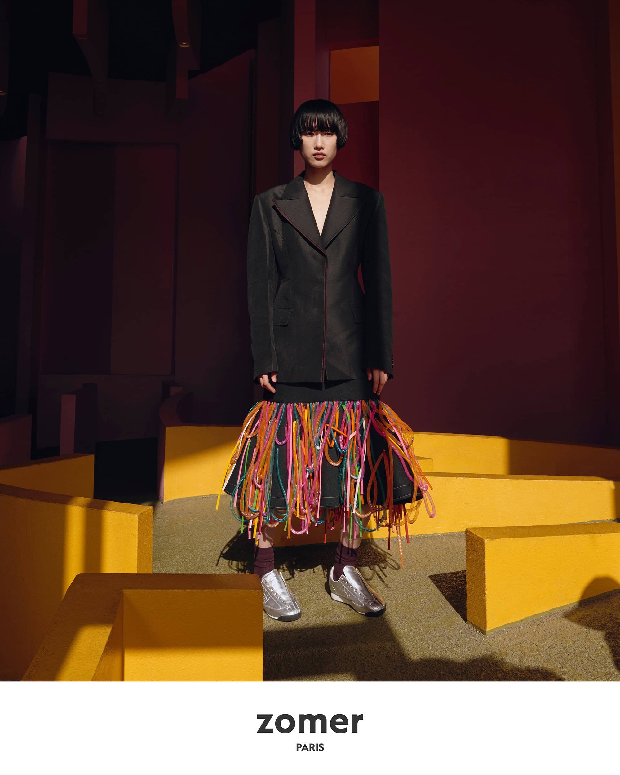 Model in a black jacket and fringed skirt standing amidst geometric yellow structures for Zomer