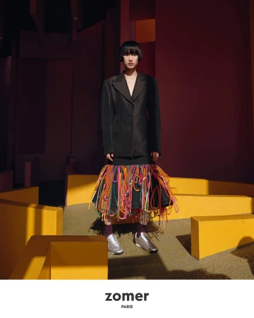 Model in a black jacket and fringed skirt standing amidst geometric yellow structures for Zomer