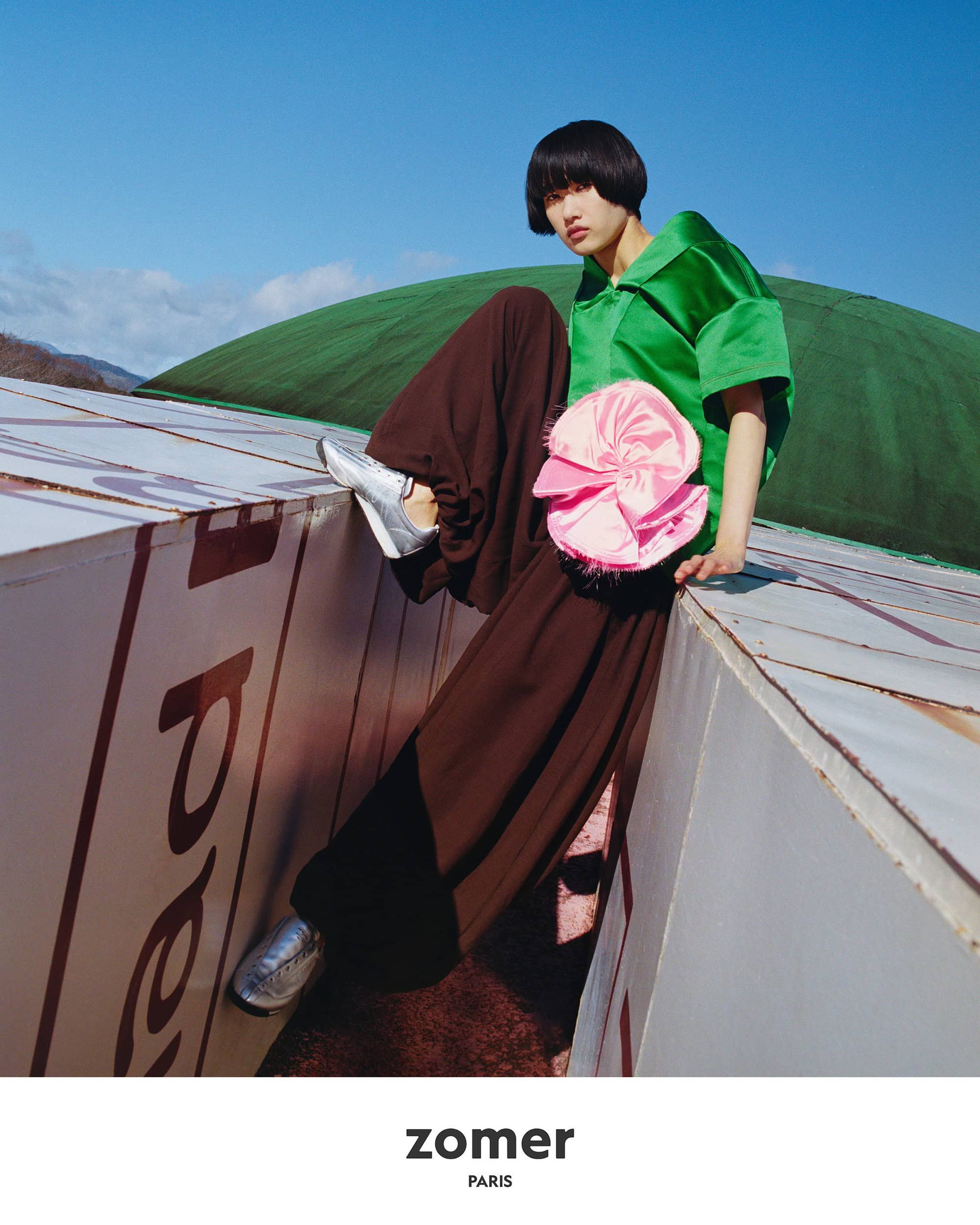 Model in green top and brown trousers posing outdoors against a large white structure and green dome under a clear blue sky