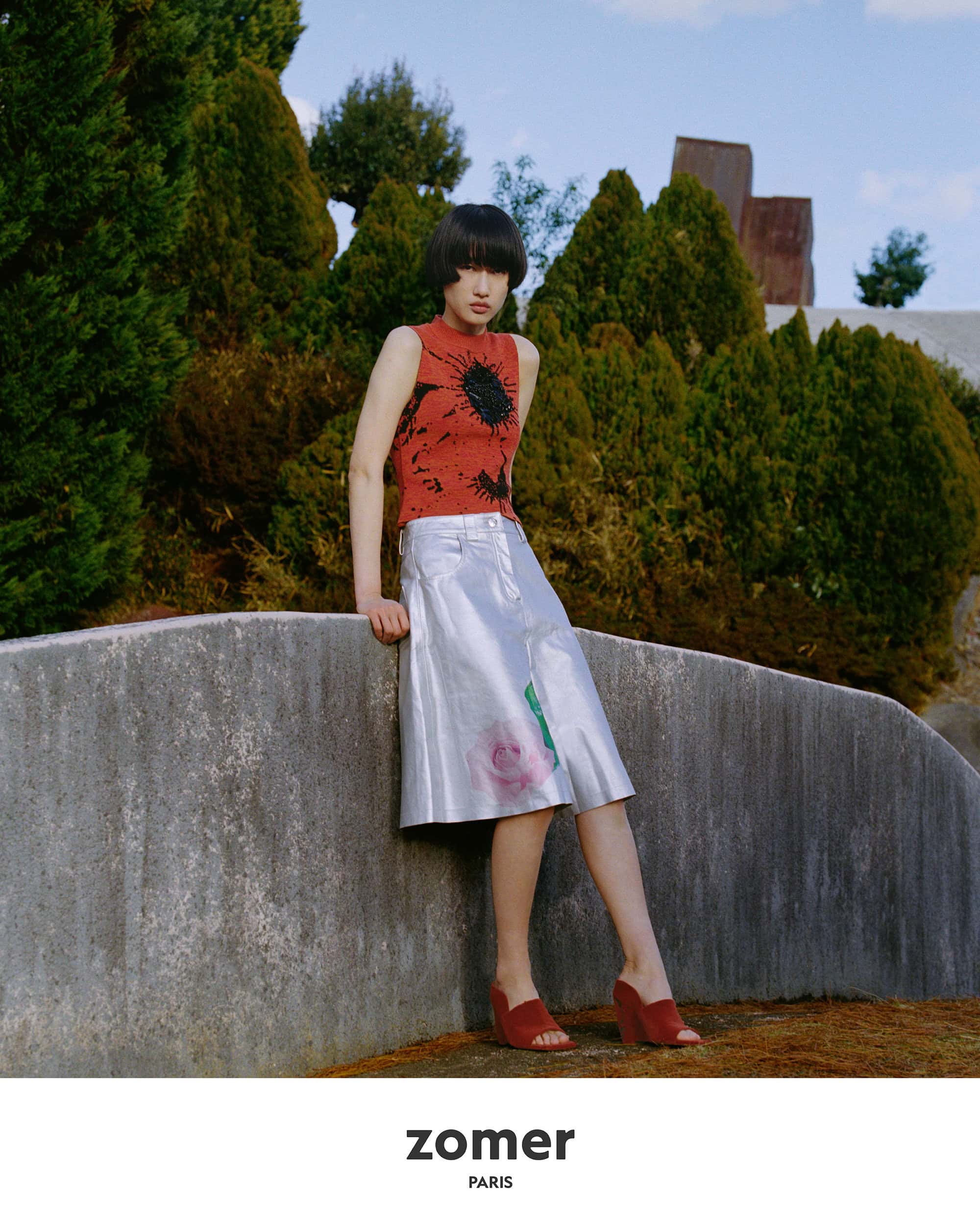 Model in a red patterned top and metallic skirt leaning against a concrete wall outdoors in Japan