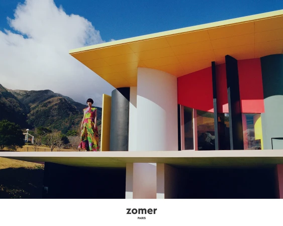 Model standing on a balcony of a brightly colored modern architectural building with mountains in the background