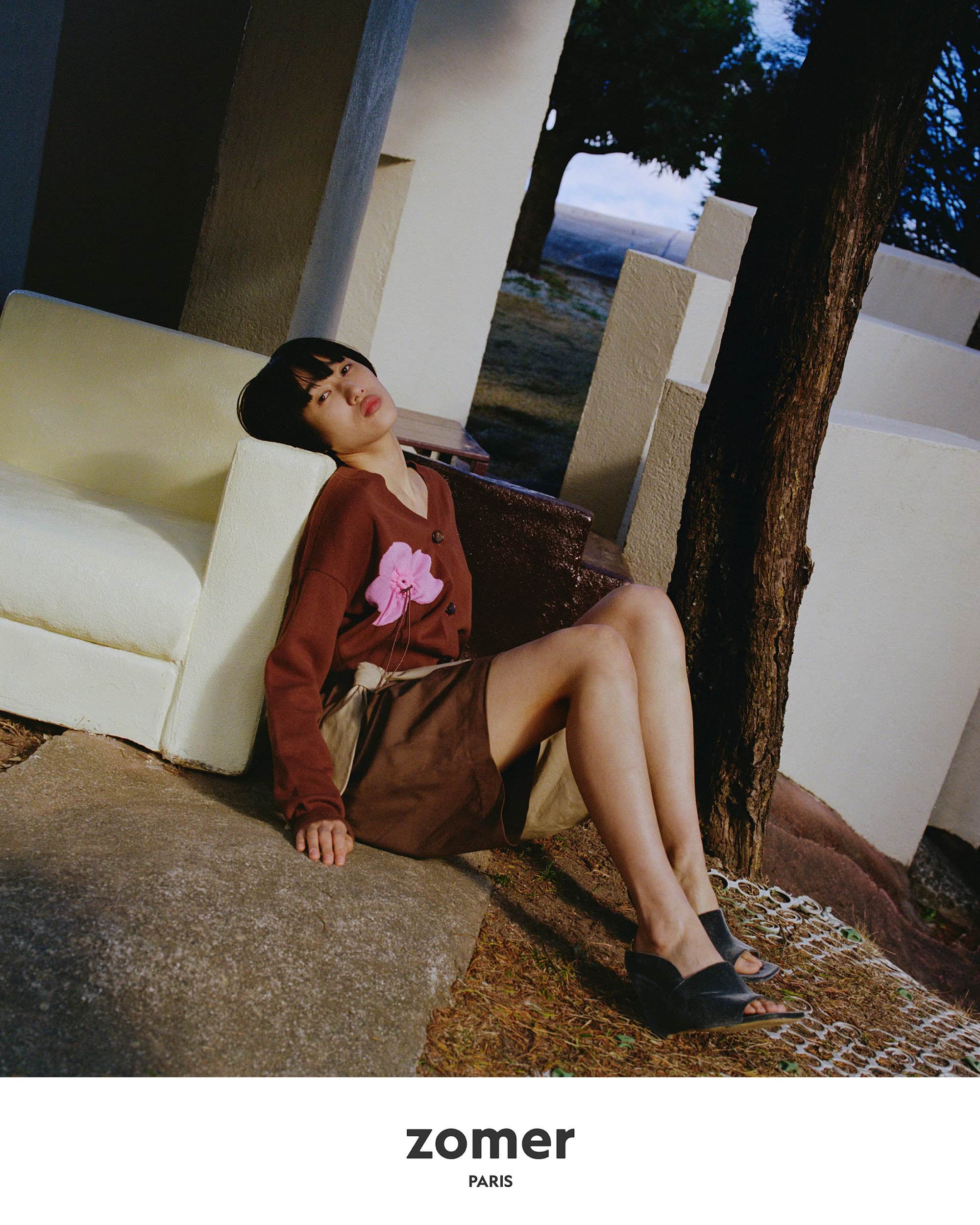 Model leaning against a light outdoor sofa wearing a brown cardigan and skirt in an architectural park setting.