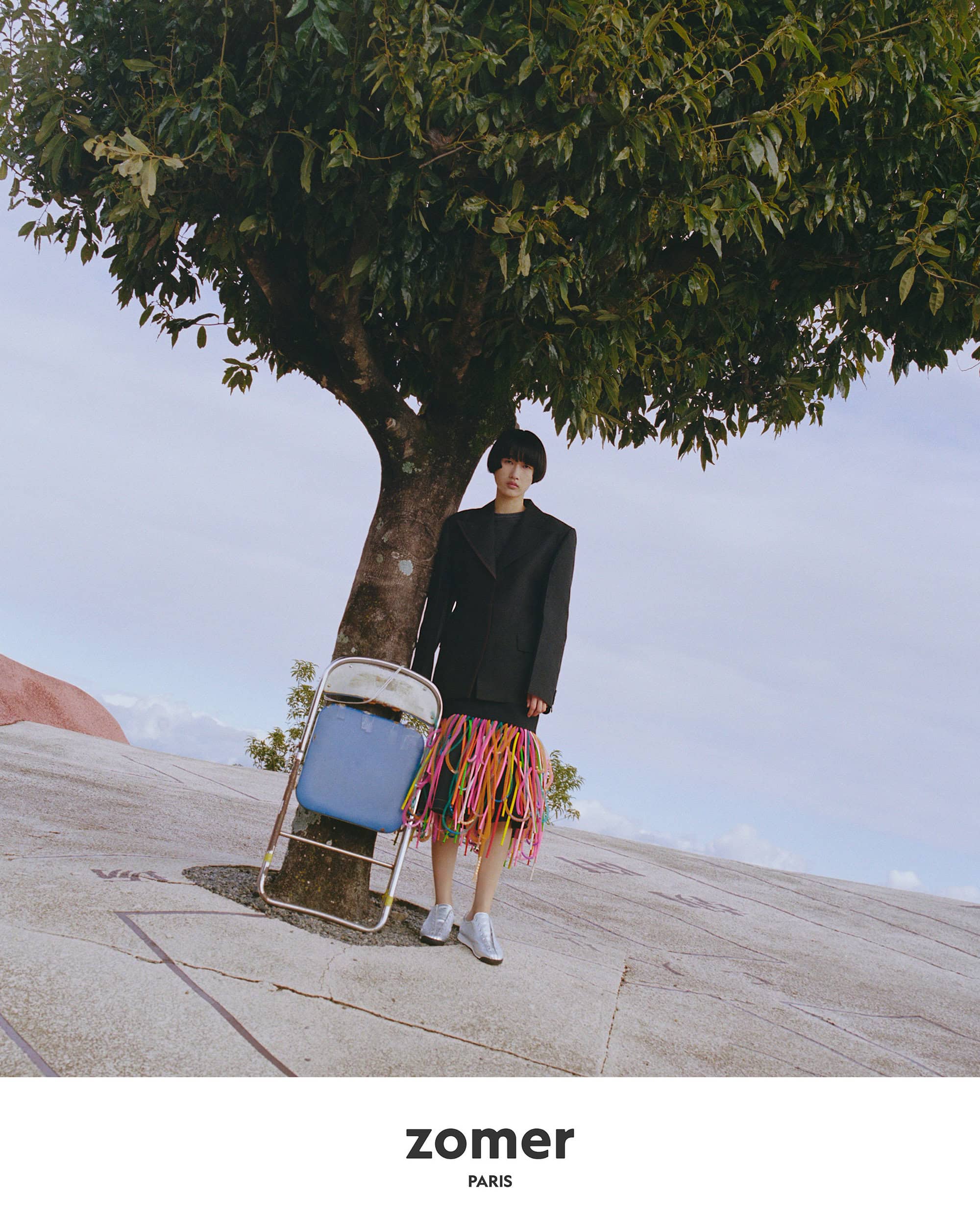 Model standing beneath a large tree on a sloped concrete ground holding a blue folding chair for Zomer Spring 2025 campaign.