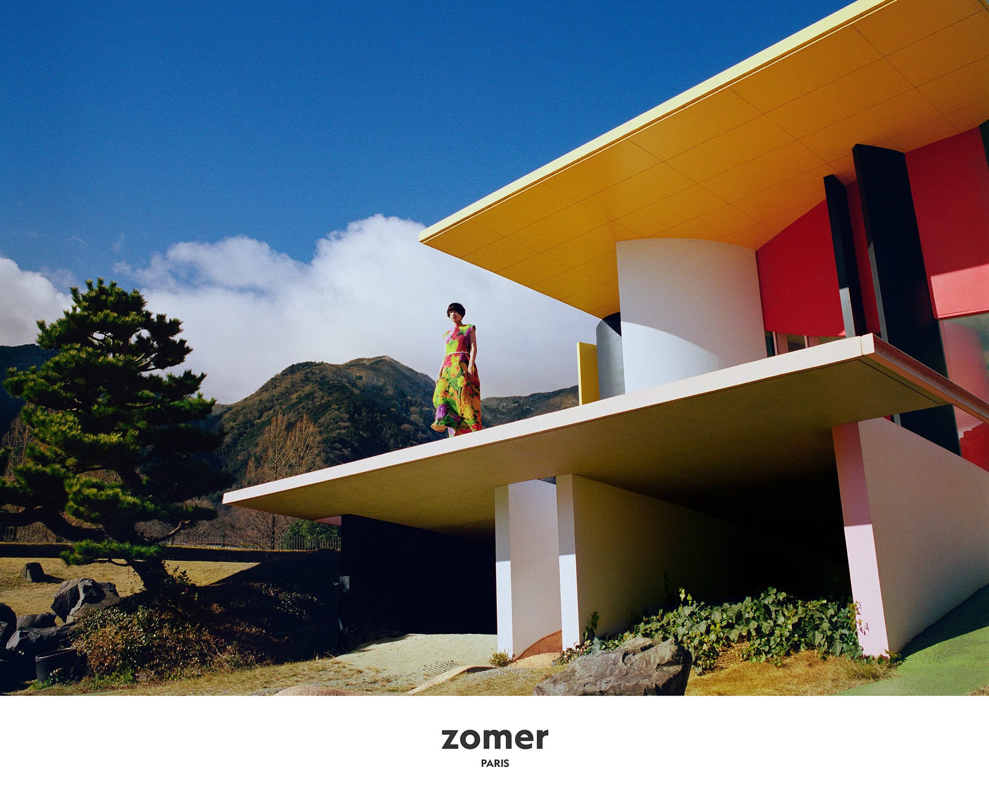 Model wearing a colourful patterned dress standing on a modern architectural balcony with mountain and sky background