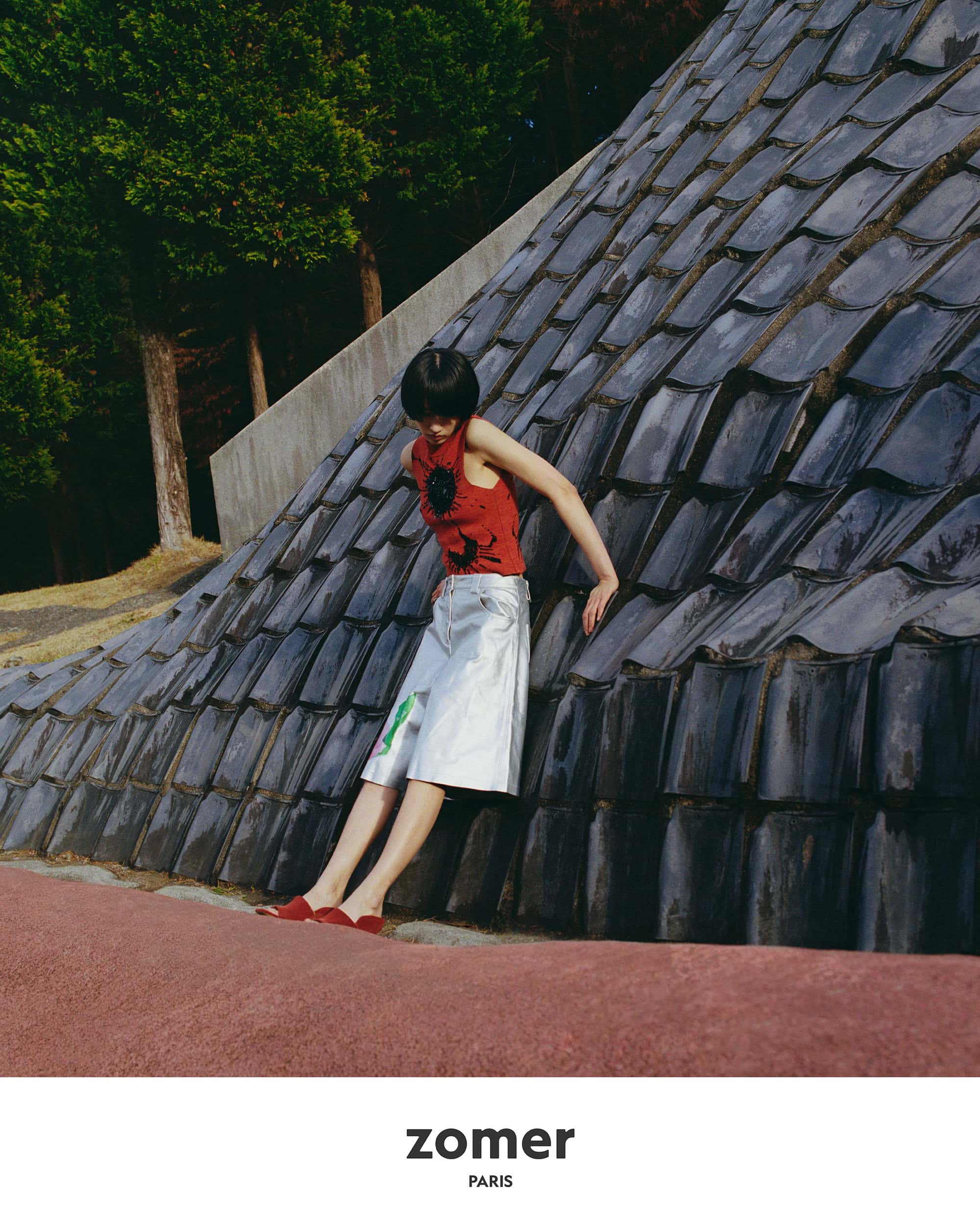 Model in red top and silver shorts leaning against a dark tiled structure in a park
