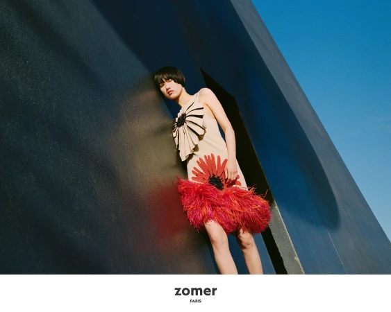 Model leaning against a dark geometric architectural wall wearing a cream dress with red floral applique and feather hem.