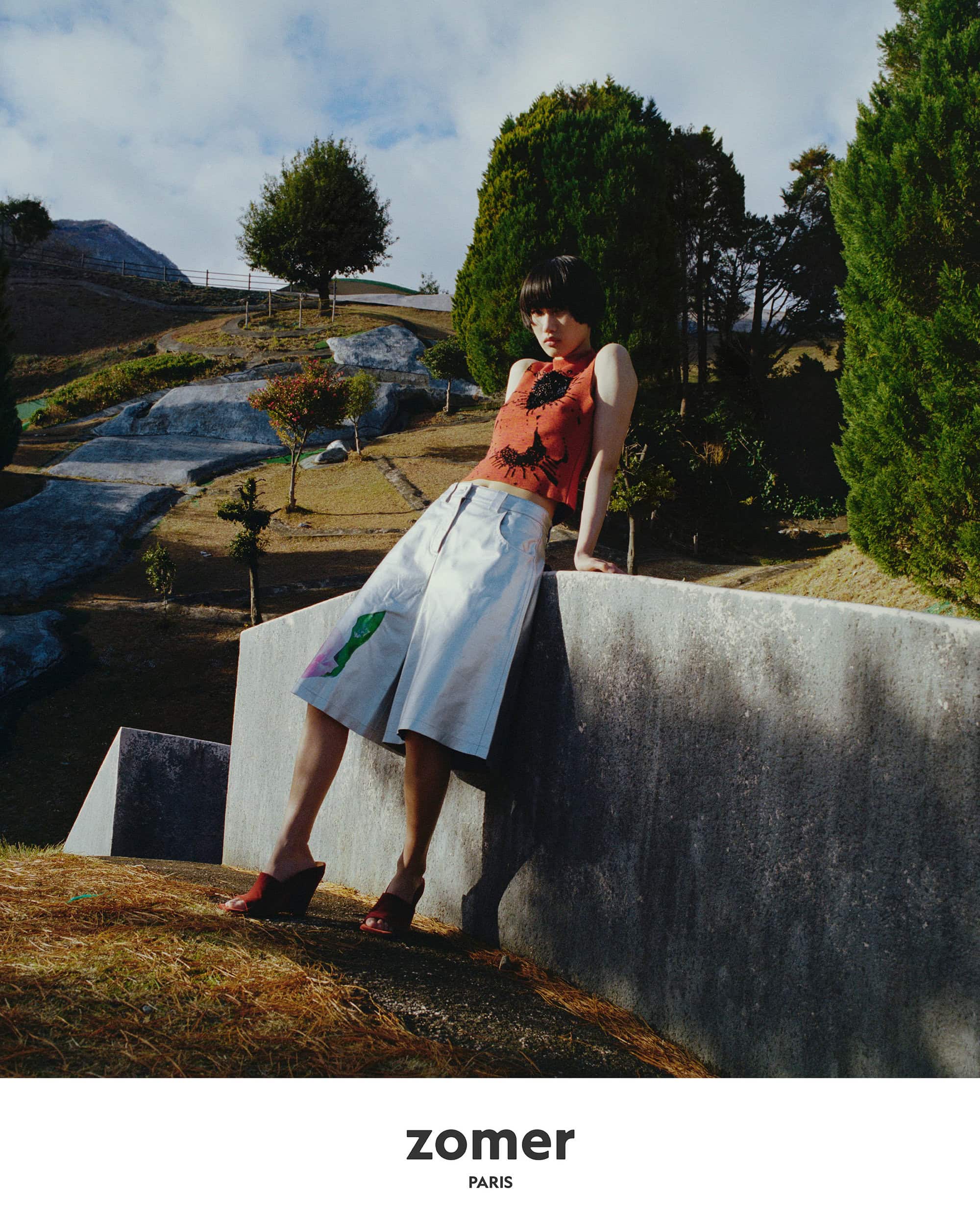Model in rust-coloured top and white shorts leaning against concrete wall in park landscape for Zomer ad campaign.