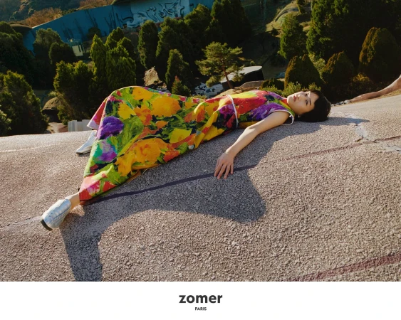 Model lying on an outdoor concrete slope wearing a colorful floral dress and white shoes amidst green trees