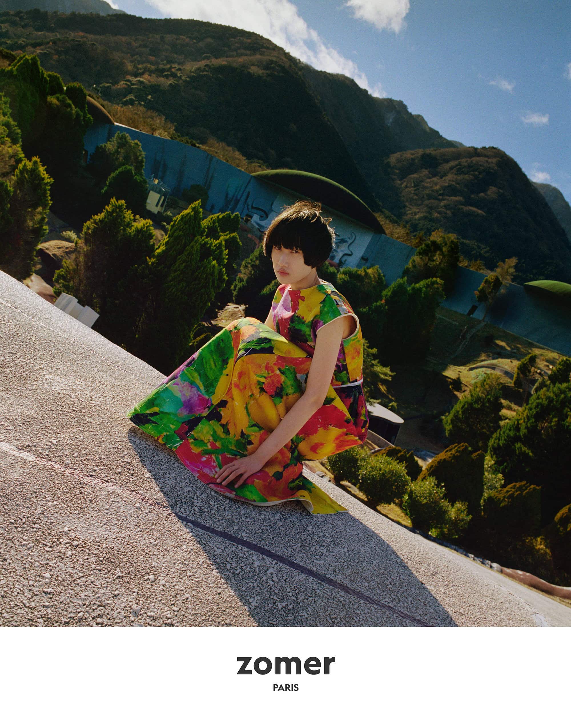 Model wearing a vibrant floral print dress crouching outdoors against a landscape of green mountains under a bright sky.